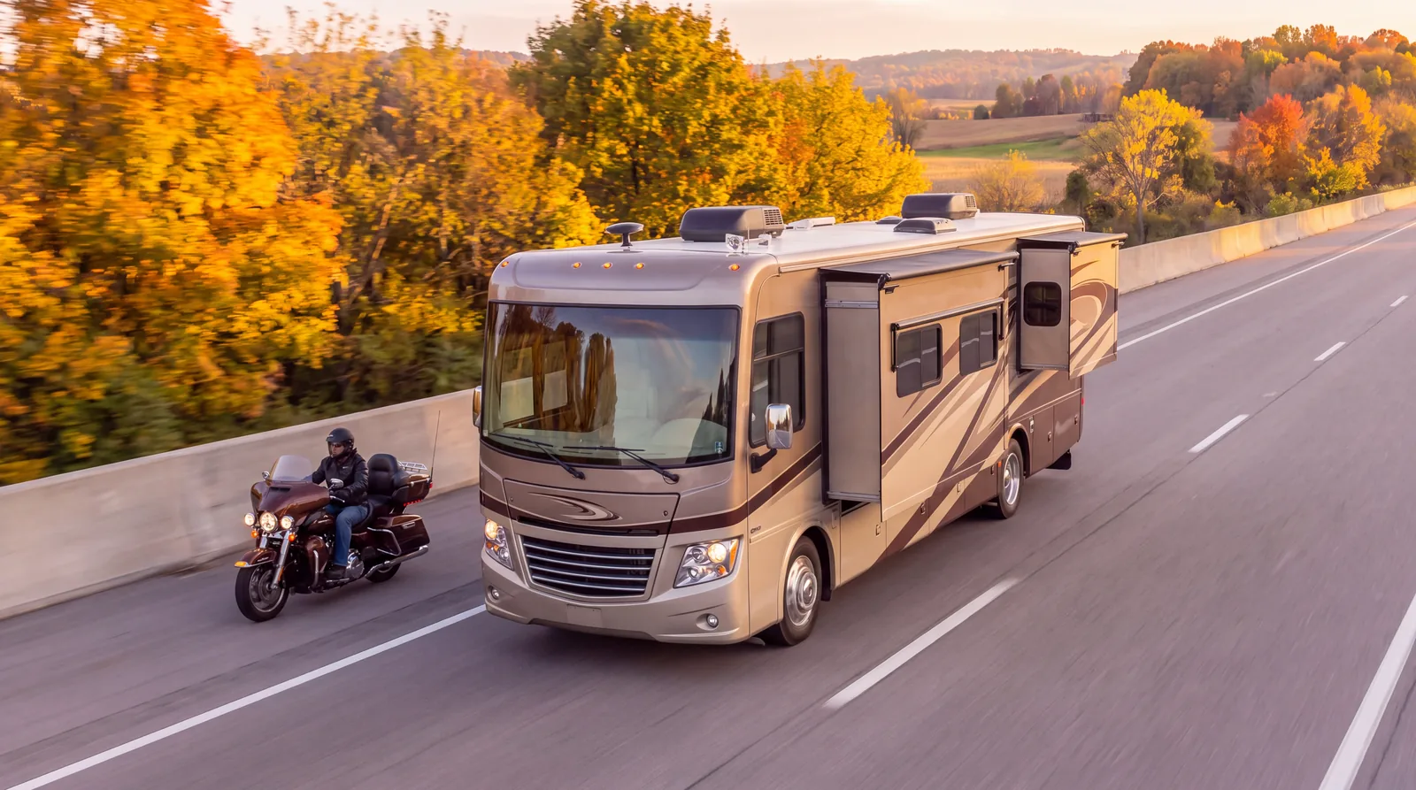 An RV and a motorcycle on a highway lined with fall trees